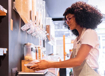 Seitenansicht einer jungen afroamerikanischen Frau, die Tasten auf einer digitalen Waage drückt, während sie eine Plastiktüte mit Schokoladen-Cornflakes während der Arbeit in einem umweltfreundlichen Lebensmittelgeschäft wiegt - ADSF38707