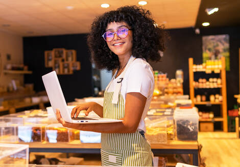 Junge lächelnde afroamerikanische Frau in Uniform und Brille mit lockigem Haar, die in die Kamera schaut und Waren in Glaskästen überprüft und einen Laptop benutzt, während sie in einem Zero-Waste-Lebensmittelgeschäft arbeitet - ADSF38711
