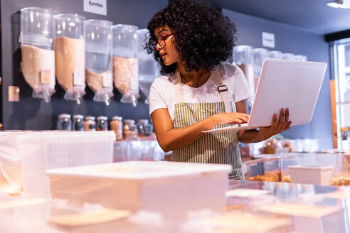 Junge afroamerikanische Frau in Uniform und Brille mit lockigem Haar, die Waren in Glaskästen überprüft und einen Laptop benutzt, während sie in einem Zero-Waste-Lebensmittelladen arbeitet - ADSF38712