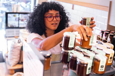 Von oben junge afroamerikanische Verkäuferin mit Brille, die Gläser mit verschiedenen Marmeladensaucen im Regal arrangiert, während sie in einem umweltfreundlichen Lebensmittelladen arbeitet - ADSF38714