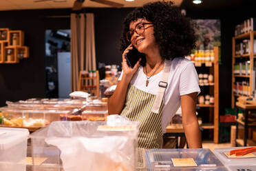 Fröhliche junge afroamerikanische Frau mit Schürze und Brille, die sich an den Tresen lehnt und mit dem Handy spricht, während sie in einem Zero-Waste-Lebensmittelgeschäft arbeitet - ADSF38721