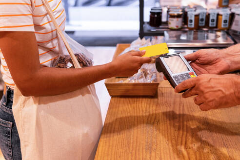Unbekannte Frau mit Tragetasche, die mit ihrer Kreditkarte für Waren an einen Verkäufer in einem Zero-Waste-Lebensmittelladen bezahlt - ADSF38728
