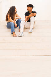 Lächelndes junges Paar in voller Länge, das sich auf einer Treppe sitzend unterhält - ADSF38735