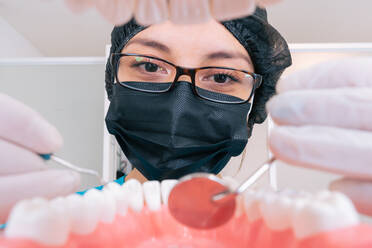 Concentrated young female medic in sterile mask gloves and eyeglasses using mouth mirror and explorer while practicing treatment on denture in dental clinic - ADSF38748