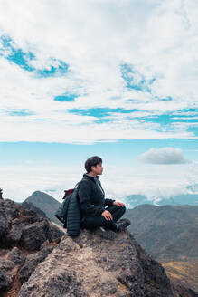 Seitenansicht einer Wanderin mit Ohrstöpseln, die auf einem Felsen in Padmasana im Hochland sitzt und der Meditation lauscht, während sie sich beim Trekking entspannt und Yoga macht - ADSF38750