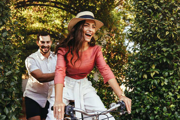 Verspielter junger Mann, der seine Freundin auf einem Fahrrad durch einen Pflanzentunnel schiebt. Glückliches junges Touristenpaar, das sich während eines romantischen Urlaubs in einem Ferienort amüsiert. Fröhlich lächelndes Paar im Freien. - JLPPF00254