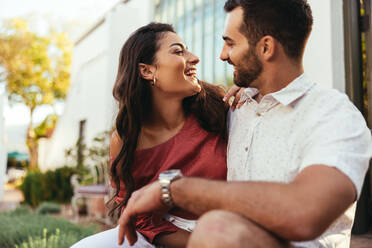 Couple laughing together while sitting outside a luxury hotel. Romantic young couple having fun together while on their honeymoon vacation. Tourist couple bonding at a holiday resort. - JLPPF00257