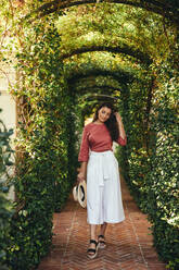 Schöne junge Touristin, die durch einen Pflanzentunnel geht. Modische junge Frau in eleganter Kleidung und mit Sommerhut. Junge Frau, die alleine an einem Urlaubsort Urlaub macht. - JLPPF00263