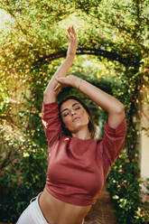 Confident young woman dancing sensually while standing alone in a plant tunnel. Beautiful young tourist woman expressing herself through dance outside a holiday resort. - JLPPF00265