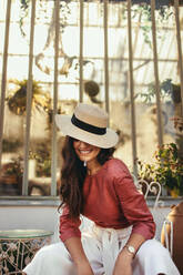 Glückliche junge Touristin, die einen Sommerhut trägt und alleine vor einem Hotel sitzt. Stilvolle junge Frau, die mit einer Blume im Mund lächelt. Frau, die alleine an einem Urlaubsort Urlaub macht. - JLPPF00273