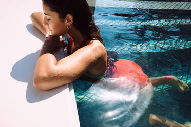 Touristin, die sich tagsüber an den Rand eines Swimmingpools lehnt. Attraktive junge Frau, die sich während eines Sommerurlaubs in einem Luxusferienort in einem Swimmingpool entspannt. - JLPPF00280