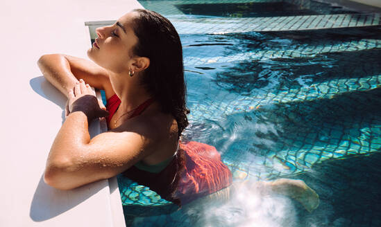 Junge Frau genießt die Sonne und lehnt sich an den Rand eines Swimmingpools. Attraktive junge Touristin entspannt sich in einem Swimmingpool während eines Sommerurlaubs in einem Luxusferienort. - JLPPF00281