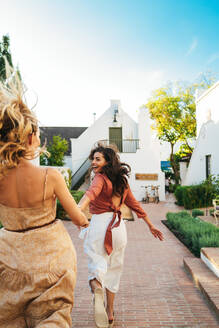 Excited young woman running while leading her friend by the hand outdoors. Happy young woman smiling at her best friend outside a holiday resort. Best friends having fun while vacationing together. - JLPPF00300