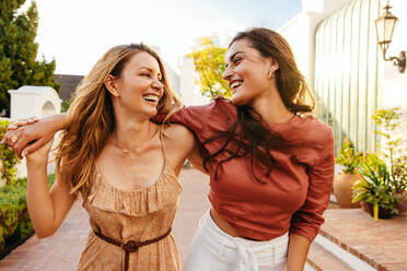 Zwei glückliche junge Frauen, die sich in den Sommerferien amüsieren. Freundinnen, die sich umarmen, während sie gemeinsam vor einem Urlaubsort spazieren gehen. Beste Freundinnen, die sich anlächeln. - JLPPF00304