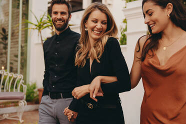 Gruppe von Freunden, die in einem Luxushotel ankommen. Drei glückliche Freunde, die fröhlich lächelnd zusammen vor einem Hotel spazieren gehen. Freunde in formeller Kleidung, die an einer gesellschaftlichen Veranstaltung teilnehmen. - JLPPF00309