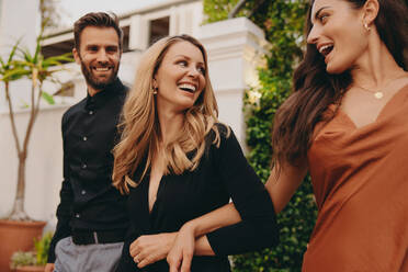 Touristengruppe von Freunden, die in einem Luxushotel ankommen. Drei glückliche Freunde, die fröhlich lächelnd zusammen vor einem Hotel spazieren gehen. Freunde in formeller Kleidung, die an einer gesellschaftlichen Veranstaltung teilnehmen. - JLPPF00310