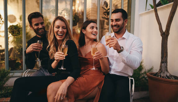 Lachende Freunde stoßen mit Weingläsern an, während sie zusammensitzen. Fröhliche Gruppe von Freunden, die vor einem Luxushotel feiern. Zwei glückliche Paare, die zusammen einen Wochenendausflug genießen. - JLPPF00328