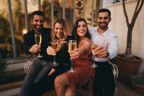 Wineglasses raised by a group of happy friends outside a luxury hotel. Four cheerful friends celebrating with wine while sitting together. Two smiling couples enjoying a weekend getaway together. - JLPPF00329