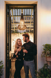 Verheiratetes Paar stößt mit Weingläsern an, während sie am Eingang eines Luxushotels stehen. Glückliches Paar, das sich fröhlich anlächelt. Paar, das einen romantischen Wochenendausflug genießt. - JLPPF00332