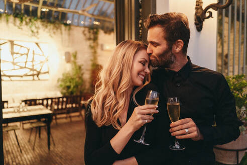 Zärtlicher Mann küsst seine Frau auf die Stirn in einem Luxushotel. Glückliches Ehepaar, das seine Liebe mit Champagnergläsern feiert. Paar, das in einem romantischen Urlaub zusammensteht. - JLPPF00336