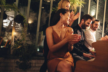 Man being applauded while playing the piano in a luxury hotel. Happy man playing the piano for his friends in the evening. Smiling group of friends enjoying a weekend getaway together. - JLPPF00345