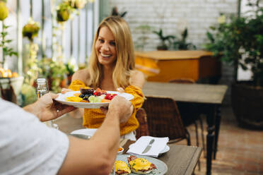 Ein gesundes Frühstück in einem gesunden Urlaub. Eine Frau lächelt, während sie von ihrem Mann einen Teller voller leckerer Früchte erhält. Ein Paar mittleren Alters genießt einen romantischen Urlaub in einem Ferienort. - JLPPF00353
