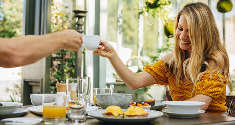 Unbeschwertes Paar stößt mit Teetassen am Frühstückstisch an. Glückliches Ehepaar beim gemeinsamen Frühstück in einem Luxushotel. Verheiratetes Paar, das einen romantischen Wochenendausflug zusammen genießt. - JLPPF00357