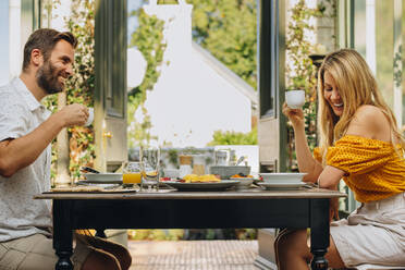 Glückliches Paar mittleren Alters, das fröhlich lächelt, während es zusammen am Esstisch Tee trinkt. Verheiratetes Paar, das einen romantischen Urlaub in einem luxuriösen Ferienort genießt. - JLPPF00358