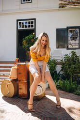 Glückliche Touristin lächelt fröhlich, während sie auf ihrem Gepäck vor einem Hotel sitzt. Weibliche Reisende wartet auf ihre Heimfahrt von einem Luxushotel. - JLPPF00377
