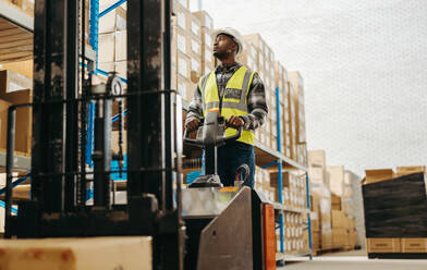 Young man in a reflective jacket driving a forklift in a large distribution warehouse. Male logistics employee moving goods around within a modern fulfilment centre. - JLPPF00388