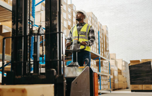 Junger Mann in einer reflektierenden Jacke, der einen Gabelstapler in einem großen Auslieferungslager fährt. Männlicher Logistikmitarbeiter, der in einem modernen Fulfillment-Center Waren umlagert. - JLPPF00388