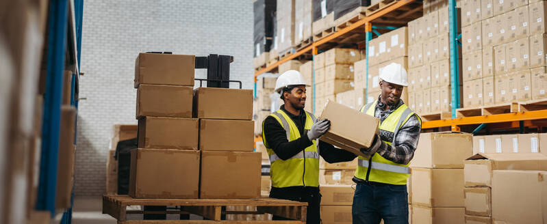 Zwei junge Männer beim Abladen von Kartons von einem Palettenhubwagen in einem großen Auslieferungslager. Zufriedene Lagermitarbeiter bei der Lagerung von Paketkartons in einem Logistikzentrum. - JLPPF00389