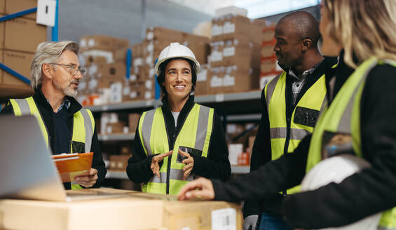 Female warehouse manager having a discussion with her team during a staff meeting. Group of multicultural logistics workers working together in a large distribution centre. - JLPPF00390