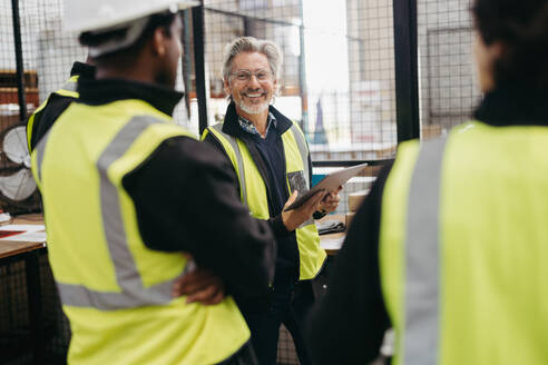 Ein älterer Lagerarbeiter lächelt fröhlich, während er eine Besprechung mit seinen Kollegen abhält. Eine Gruppe multikultureller Logistikmitarbeiter arbeitet als Team in einem großen Auslieferungslager. - JLPPF00392