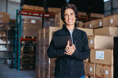 Portrait of a warehouse supervisor smiling at the camera while holding a digital tablet in her hand. Happy mature woman working with warehouse management software in a distribution centre. - JLPPF00396