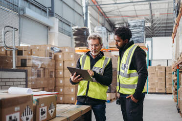 Two warehouse workers using a digital tablet while recording inventory. Logistics employees working with warehouse management software in a large distribution centre. - JLPPF00401
