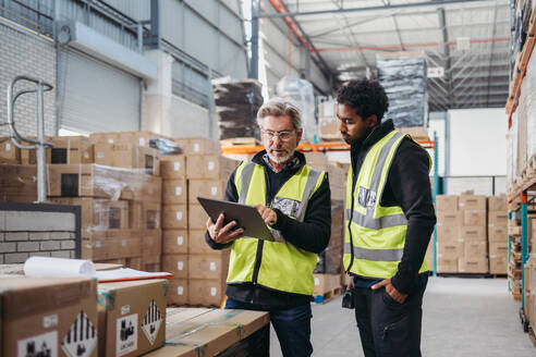 Zwei Lagerarbeiter, die ein digitales Tablet verwenden, während sie das Inventar erfassen. Logistikmitarbeiter bei der Arbeit mit einer Lagerverwaltungssoftware in einem großen Vertriebszentrum. - JLPPF00401