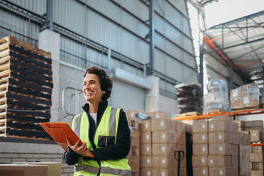 Mature warehouse manager looking away with a happy smile while holding a clipboard. Cheerful woman working in a large distribution centre. - JLPPF00402