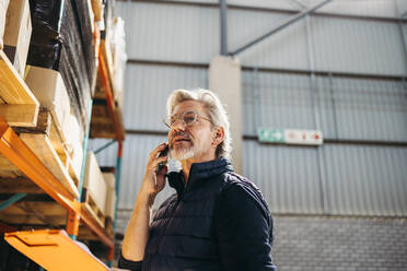 Senior man speaking on a phone call while standing in a distribution warehouse. Senior logistics manager making shipment plans over the phone in a large fulfillment centre. - JLPPF00404