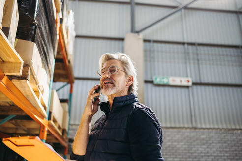 Ein älterer Mann führt ein Telefongespräch, während er in einem Auslieferungslager steht. Ein leitender Logistikmanager, der in einem großen Logistikzentrum am Telefon Versandpläne erstellt. - JLPPF00404