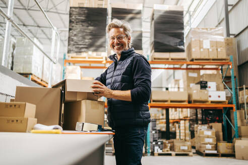 Fröhlicher älterer Mann, der in die Kamera lächelt, während er Kartons in einem Auslieferungslager packt. Fröhlicher Lagerarbeiter, der in einem großen Logistikzentrum Waren für den Versand vorbereitet. - JLPPF00405