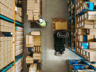 Top view of a warehouse worker moving package boxes in a large logistics centre. Aerial view of a young man transporting goods with a pallet truck within a modern fulfilment centre. - JLPPF00417