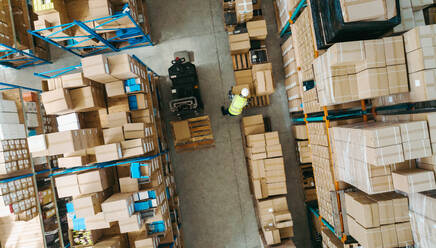 High angle view of a warehouse employee moving goods in a large logistics centre. Aerial view of a young man transporting package boxes with a pallet truck within a modern fulfillment centre. - JLPPF00420