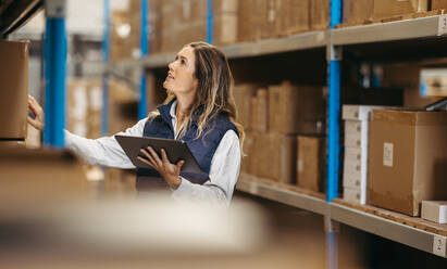 Female warehouse manager counting stock while holding a digital tablet. Woman doing inventory control using warehouse management software in a modern distribution centre. - JLPPF00429