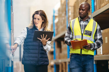 Eine Lagerverwalterin zeigt auf ein Paket in einem Regal, während sie sich mit ihrem Kollegen unterhält. Zwei Logistikmitarbeiter bei der Bestandskontrolle in einem großen Vertriebszentrum. - JLPPF00432