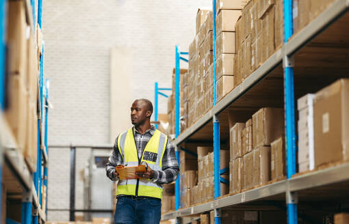 Warehouse supervisor taking inventory using a clipboard and a barcode reader in a modern logistics centre. Young black man looking at parcel boxes while standing in between storage shelves. - JLPPF00435