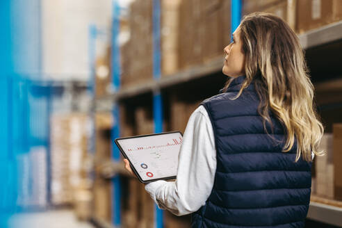 Rearview of a warehouse manager using warehouse management software on a digital tablet. Female logistics employee working with statistical reports on her inventory management dashboard. - JLPPF00438