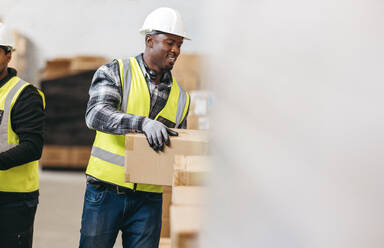 Happy young man moving cardboard boxes while working with a colleague in a warehouse. Male logistics worker storing package boxes in a large distribution centre. - JLPPF00442