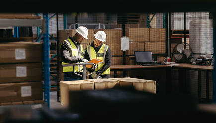 Lagerarbeiter lesen ein Klemmbrett, während sie in einem Logistikzentrum mit einem gepackten Palettenhubwagen stehen. Zwei junge Männer nehmen in einem großen Verteilungszentrum Pakete für den Versand entgegen. - JLPPF00445