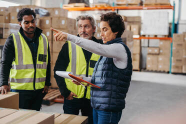 Warehouse manager reading a document while having a discussion with her team during a meeting. Group of diverse employees working together in a large distribution centre. - JLPPF00452